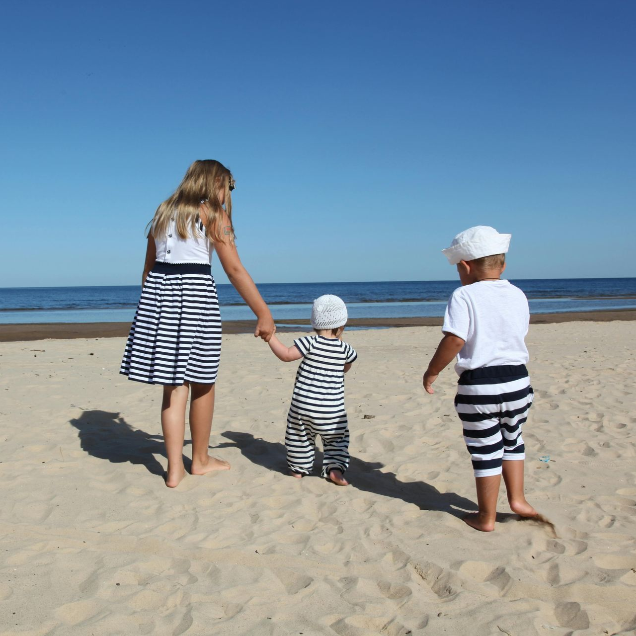 Three children holding hands walk barefoot on a sunny beach toward the ocean.