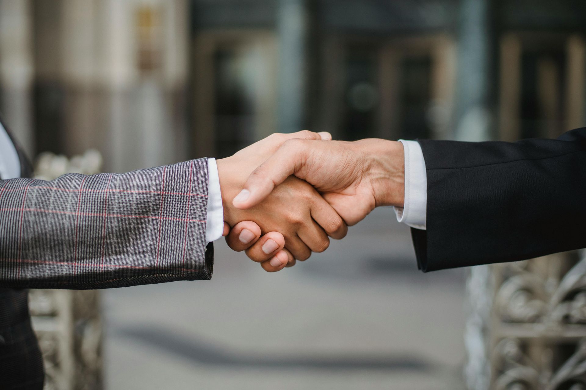 Two people in business attire shaking hands outdoors with a blurred building in the background