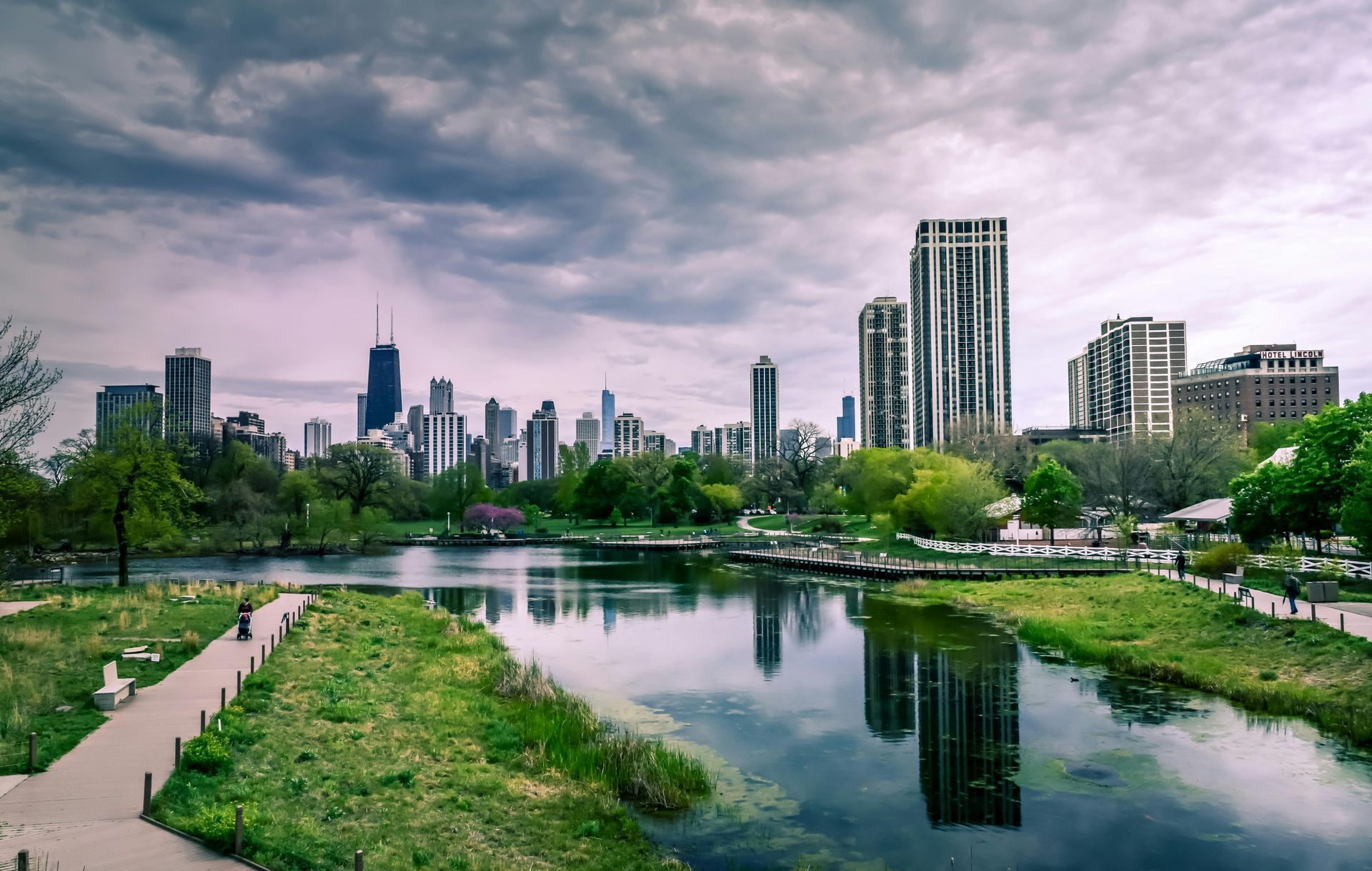 City skyline reflected in a calm river beside a green park under a cloudy sky
