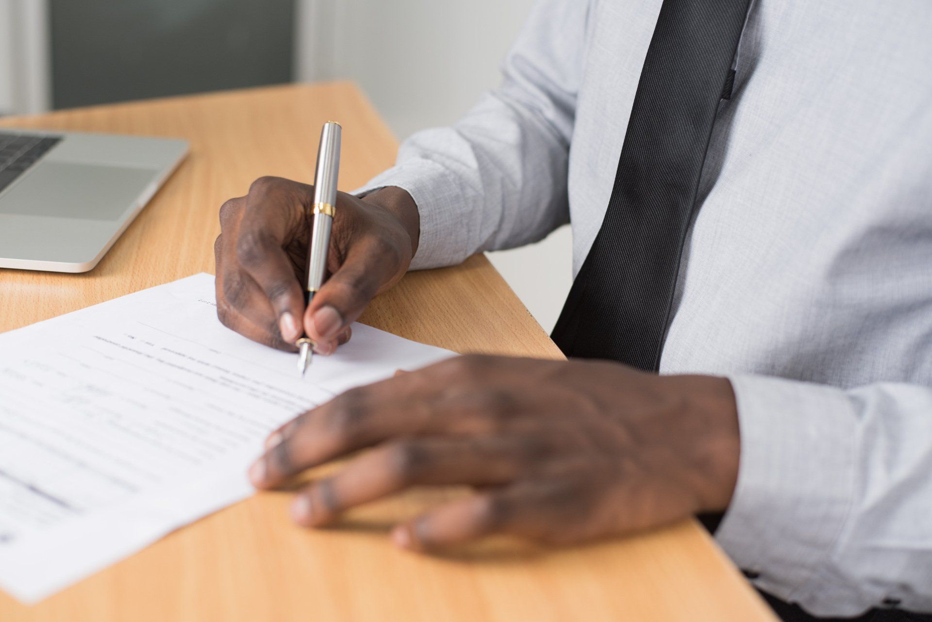 Hands writing with a pen on paper at a desk beside a laptop
